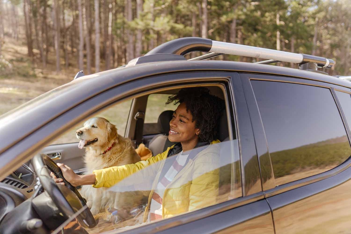 Leende svart kvinna i gul jacka kör svart bil i naturen, med golden retriever i passagerarsätet.