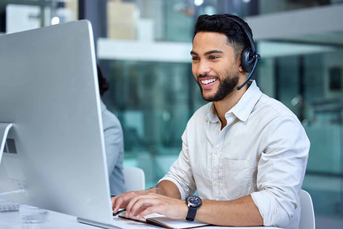 Ung mörkhårig snygg man arbetar i kundtjänst framför en datorskärm och med headset.